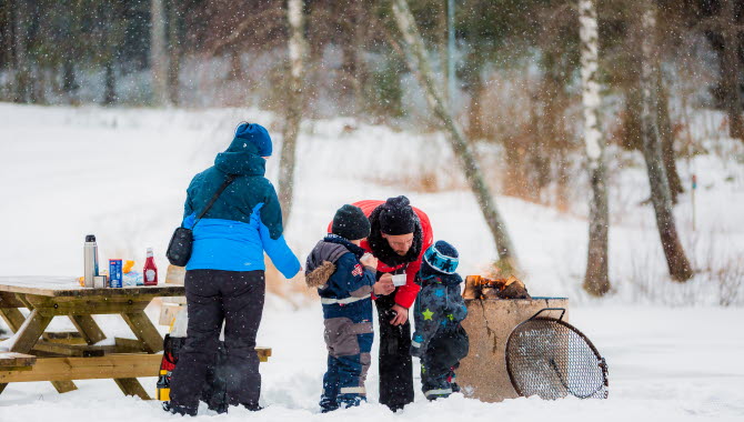 En familj med två barn grillar korv vid en grillplats. Det är vinter med snö.