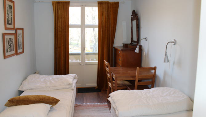 Ett rum med två sängar, ett litet bord med två stolar, en byrå och ett fönster med gardiner.