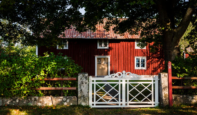 Ett rött hus med vita knutar och gul vacker dörr står omgivet av en lummig trädgård som väntar innanför en vit grind med snickarglädje på toppen. 