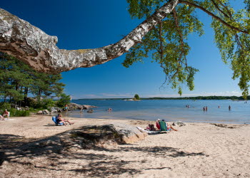 Utsikt över sandstranden där personer solar vid Vänerns vatten.