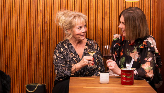 Two women are holding a wine glass. One woman is blonde and has updo hair. The other woman has brown shoulder-length hair. Both women look at each other. Both women wear dark blouses. The wall behind them consists of a light wood panel.