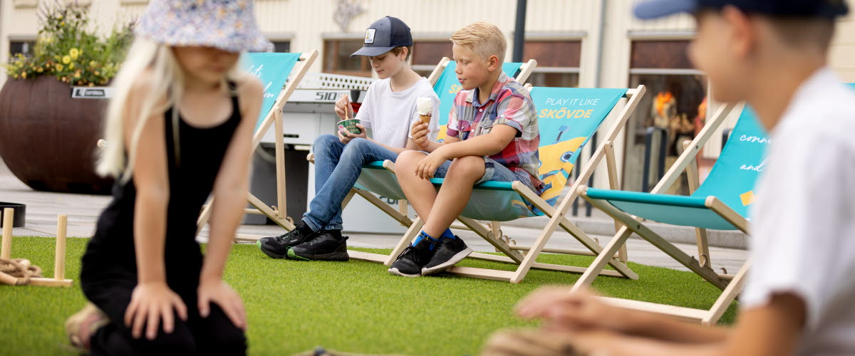 Två barn spelar ett spel på gräset medan två killar sitter i varsin brassestol i bakgrunden.