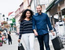 A man and woman with wide smiles walk along a shopping street, both carrying a bag in hand.