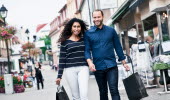 A man and woman with wide smiles walk along a shopping street, both carrying a bag in hand.