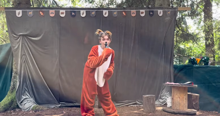 En person i rävdräkt står ensam framför scenen med mikrofon i hand, mitt i ett uttrycksfullt framträdande.