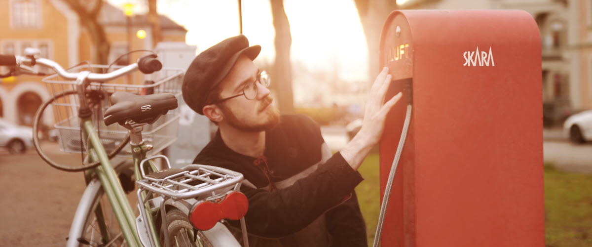Man med glasögon och keps som pumpar sin cykel vid en stor röd cykelpump i plåt. 