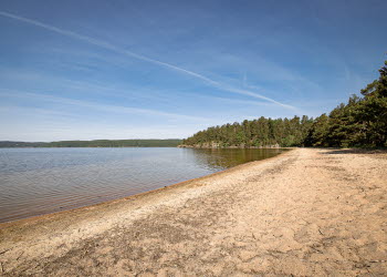 En bild på en lång och bred sandstrand som breder ut sig intill en sjö med klart vatten. Himlen är bra och i bakgrunden skymtas skog.