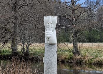 En skulptur in sten som föreställer ett ansikte som håller ett finger över munnen. Runt omkring är det gräs, träd och vatten.
