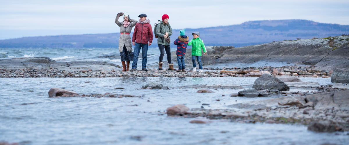 Tre vuxna och två barn är ute och vandrar. Bakom dem syns berget Kinnekulle och framför dem syns sjön Vänern. 