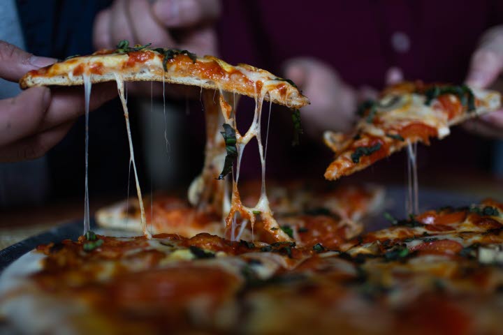 En närbild på händer som plockar varsin pizza slice från en pizza. Det är gott om osttrådar.