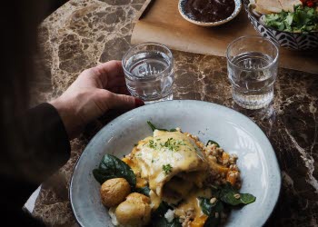 Någon sitter och äter vid ett runt bord och håller i ett dricksglas. En tallrik med mat och tillbehör vid sidan om.
