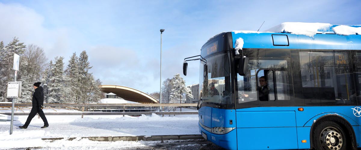 Framdelen av en blå bussar, i bakgrunden ser du snö och skog, en människa går ut ur bilden till vänster.