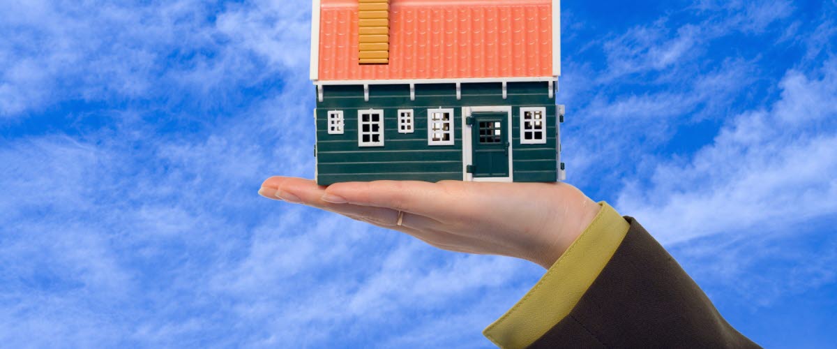 Ett litet miniatyr hus hålls upp i en hand med blå molnbeklädd himmel i bakgrunden.