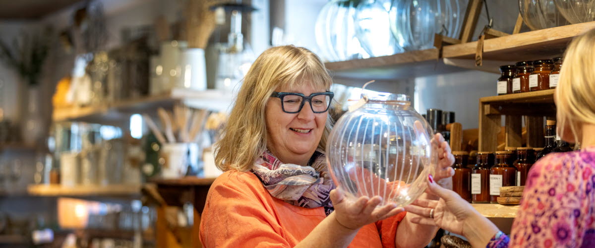 En kvinna i svarta glasögon och korallfärgad tröja står och tittar på en glasvas i en butik. 