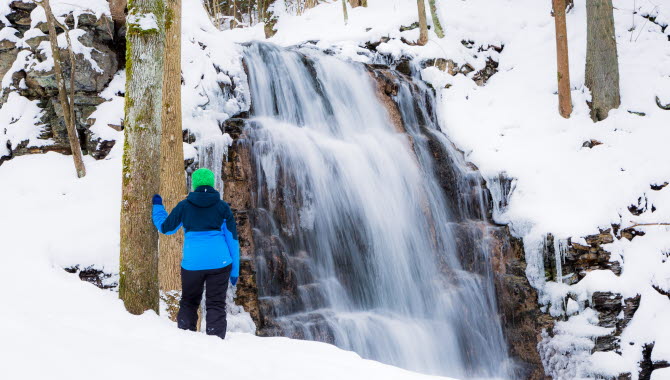 Ett vattenfall i ett snöigt landskap. Framför fallet står en person i blå jacka och tittar på det forsande vattnet.