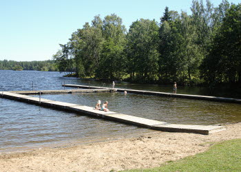 En bild på en sjö omgiven av skog. I vattnet ligger en träbrygga och några människor står på den. Intill sjön är en smal sandstrand som övergår i grönt gräs.