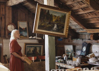 En kvinna i röd klänning är på en loppis i en lada. På en bärande stolpe hänger en sned tavla med guldram.