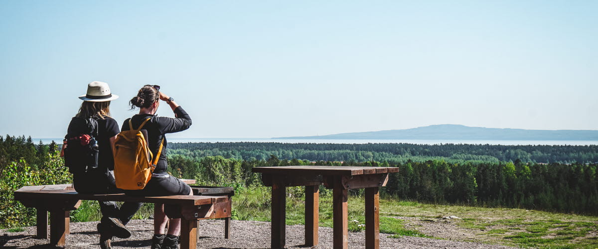 Två kvinnor sitter på en bänk och blickar ut över landskapet med Kinnekulle i horisonten.