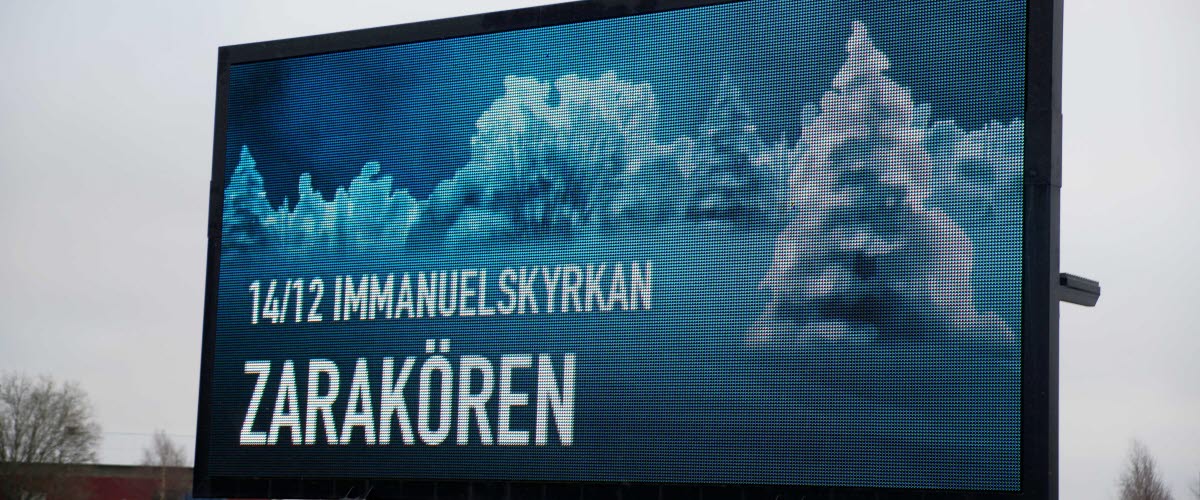 Stor digital skylt som står intill vägen visar reklam för Zarakörens konsert, mot en vintrigt landskap.