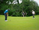 Bilden visar en golfgreen med fyra golfare och skog i bakgrunden. 
