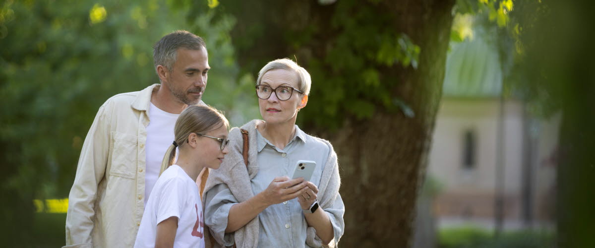 En man, en kvinna och en flicka står i en park och tittar ned i en telefon.