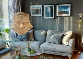 En bild på en soffa med prydnadskuddar som står mot en mörk vägg med tavlor på. Framför soffan står två runda bord. Solen lyser in genom ett fönster. Från taket hänger en stor taklampa.