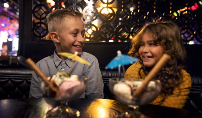 Två barn, en kille och en tjej, sitter på restaurang med två festliga efterrätter framför sig. 