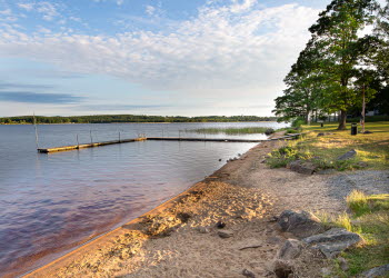 En bild på en smal sandstrand som kantas med stenbumlingar och lite växtlighet. Intill stranden ligger en sjö med en brygga i.