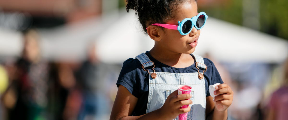 Liten flicka med svart krulligt hår iklädd snickarbyxor och coola solglasögon. I handen har hon en rosa glass. 