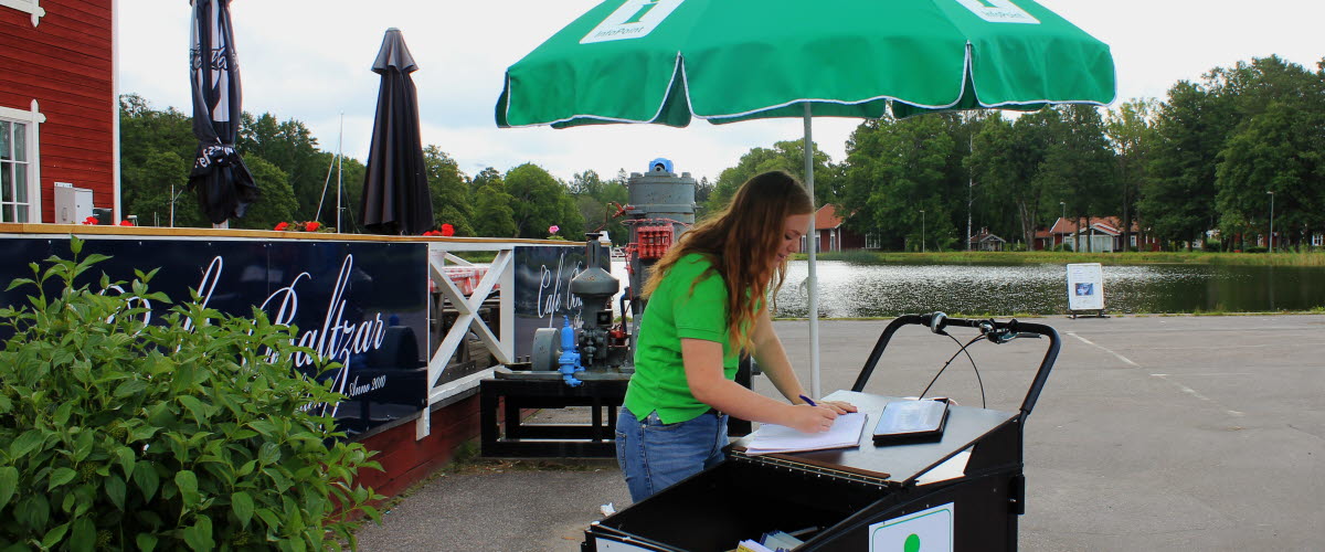 En packcykel med broschyrer, ett grönt parasoll och en kvinna i grön tröja står utanför en uteservering vid Göta kanal.