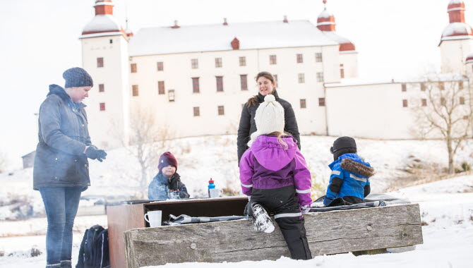 Vuxna och barn vid en grillplats med Läckö Slott i bakgrunden. På marken ligger snö.