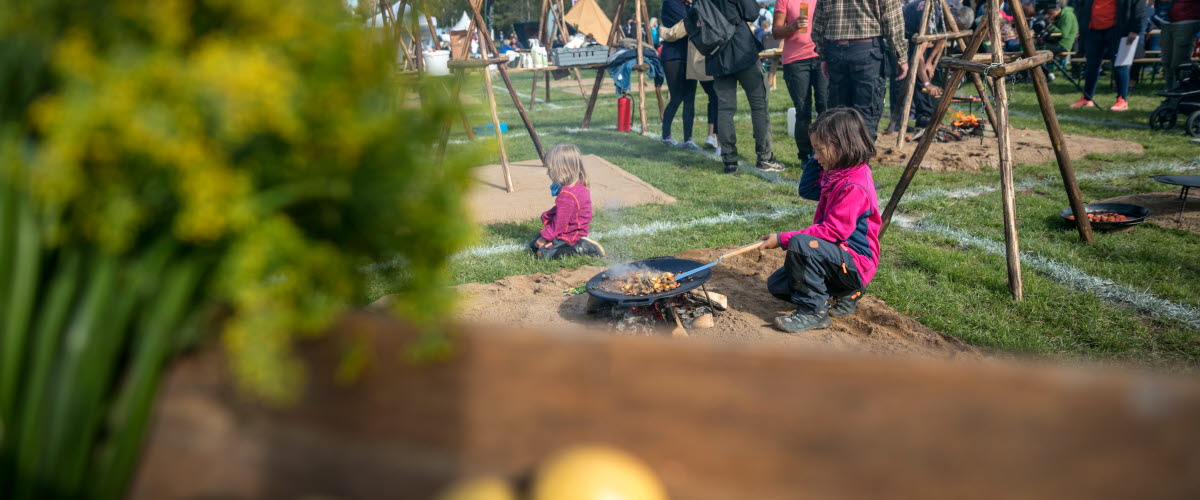 Två små flickor i rosa fleecetröjor lagar mat på en murikka på ett festivalområde bland massor av människor på en utomhusfestival