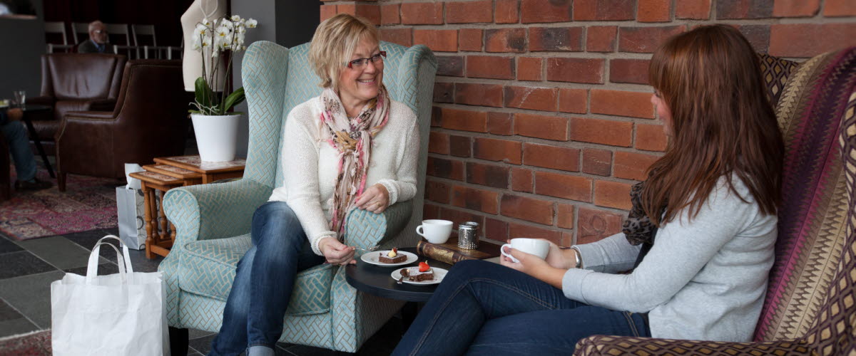 Two women are sitting in two armchairs, talking and drinking some coffee. 