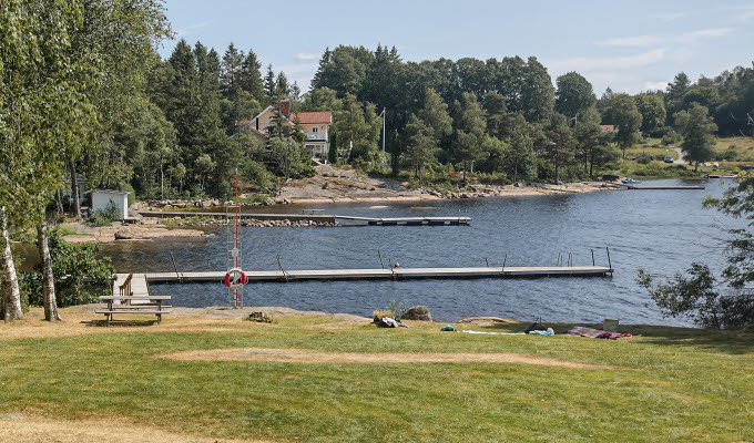 Intill en stor gräsmatta ligger en sjö med härligt blått vatten. I sjön ligger en brygga. I bakgrunden skymtas skog och hus.
