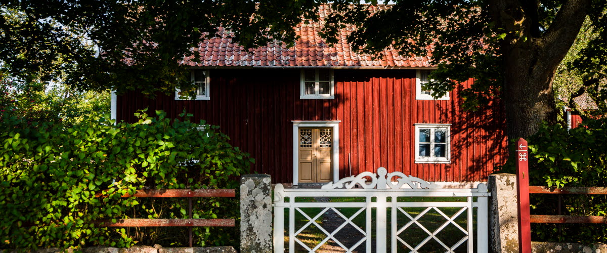 Ett rött hus med vita knutar och gul vacker dörr står omgivet av en lummig trädgård som väntar innanför en vit grind med snickarglädje på toppen. 