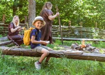 Pojke med gul ryggsäck sitter och bakar ett bröd. i bakgrunden syns en eld och två stenåldersklädda människor.
