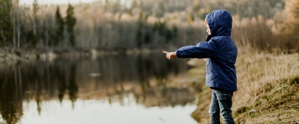 Ett barn i blå jacka och ett par gummistövlar pekar ut över vatten. Bakgrunden i fylld av växtligheter. 