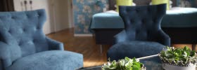 Hotelroom with double bed in the back and two blue armchairs in front. In the foreground is a table with two flowerpots.