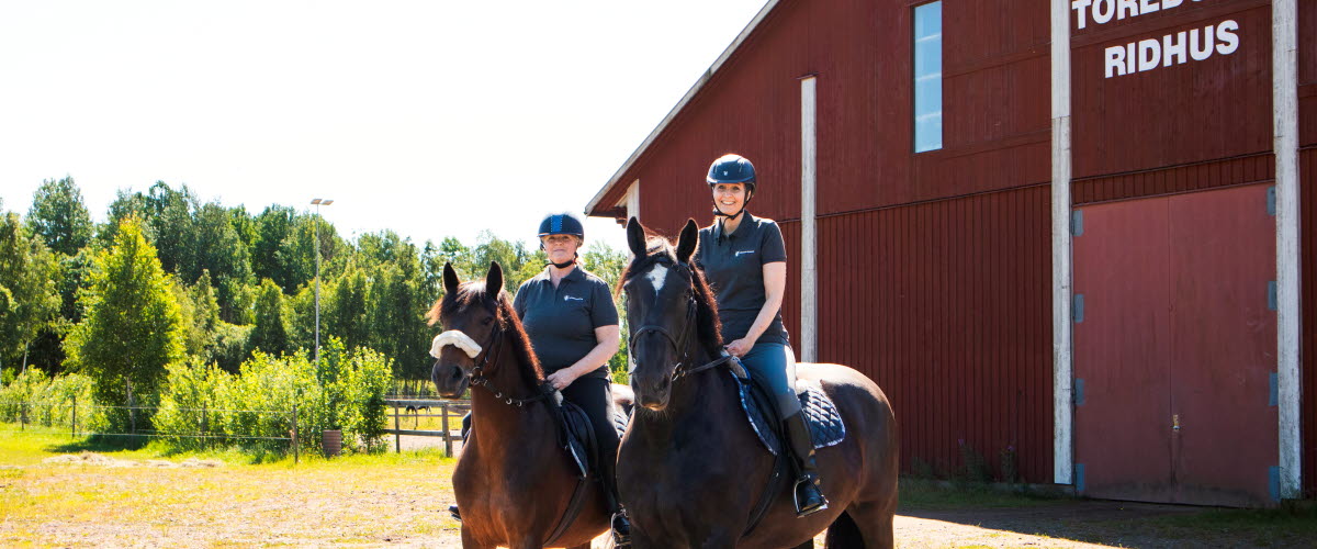 Två kvinnor rider utomhus. I Bakgrunden syns en stor röd byggnad med texten Töreboda ridhus.