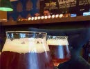 Två glas öl och en skål med nötter på ett bord med bardisk i bakgrunden.