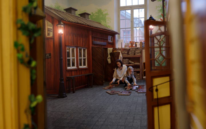 En vuxen och ett barn sitter på golvet framför ett äldre rödmålat hus i miniatyr inne i en utställning. 