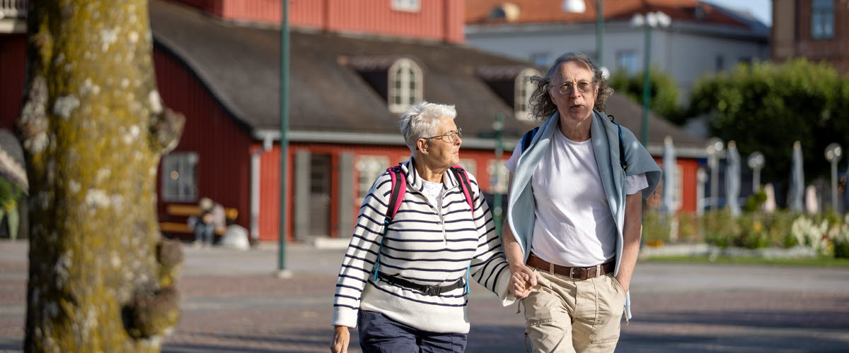 Ett äldre par håller varandra i handen på ett torg med ett rött hus i bakgrunden.