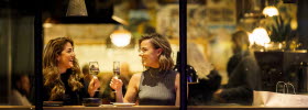 Two women are sitting at a table placed by a window. They each raise a glass toward each other for a toast.