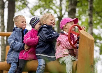 Fyra små barn sitter bakom varandra på en lång trästock. Barnet längst fram har en ratt på en trästolpe framför sig. 