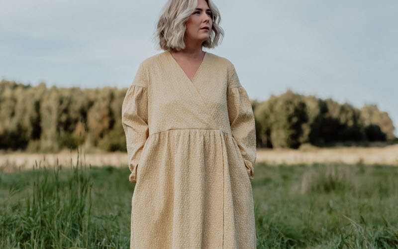 Kvinna med blont, axellångt hår står på en grön äng iförd en lång, ljusgul klänning med lång ärm. I bakgrunden syns skog och en klar himmel.