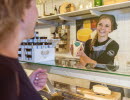 Woman standing behind a cheese counter and holding up a cheese for a customer.