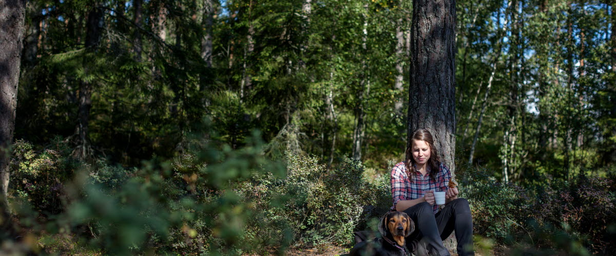 Flicka med hund sitter och vilar mot ett träd i skogen.