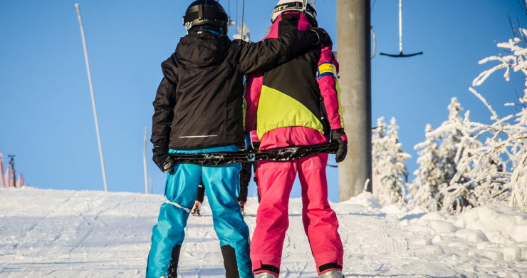 Två barn i färgglada kläder påväg upp för backen i ankarliften omgivna av vinterlandskapet. 