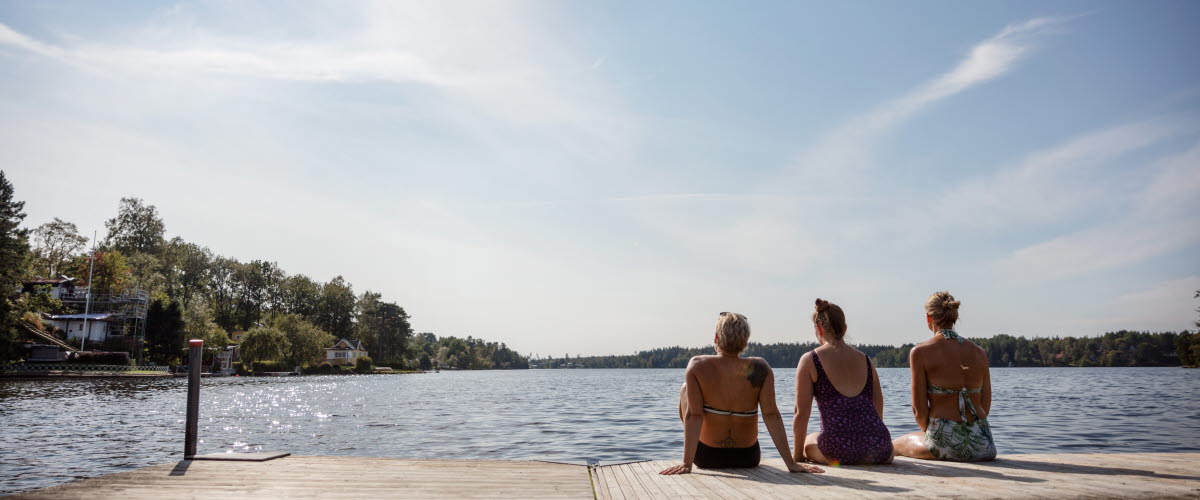 Tre kvinnor sitter på en brygga med ryggen mot kameran. Sjön i bakgrunden.