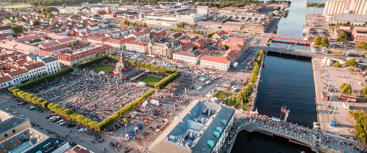 Flygbild över Lidköpings centrum. Till vänster är torget, fullt med folk. Till höger rinner ån Lidan som mynnar ut i Vänern längst bort i bild. Två broar korsar ån.
